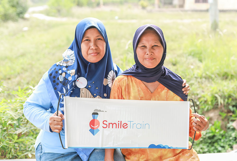 Endang smiling and holding a Smile Train banner with an older Smile Train patient