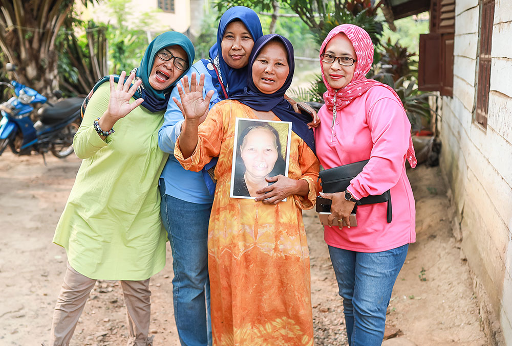 Endang smiling with an older Smile Train patient and her family and holding a photo of her before cleft surgery